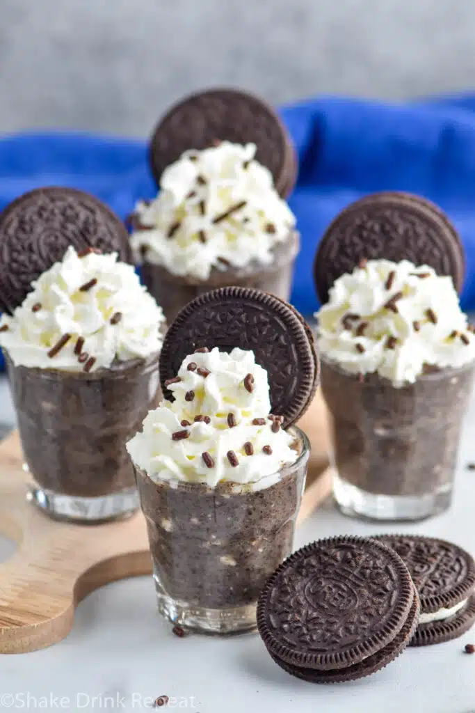 Photo of Cookies and Cream Pudding Shots garnished with whipped cream, sprinkles, and Oreo cookies. Extra Oreo cookies on counter for garnish.