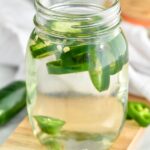 Close up photo of a jar of Jalapeno Infused Vodka recipe. Extra sliced jalapeno peppers beside jar.