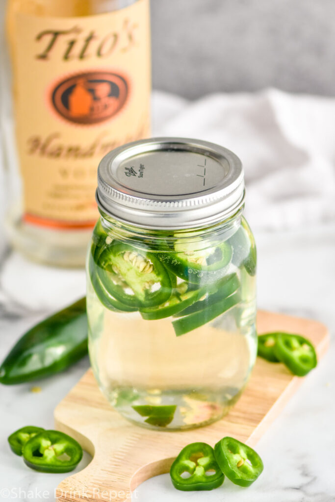 Overhead photo of a jar of Jalapeno Infused Vodka recipe with extra jalapeno peppers and a bottle of Tito's vodka in the background.