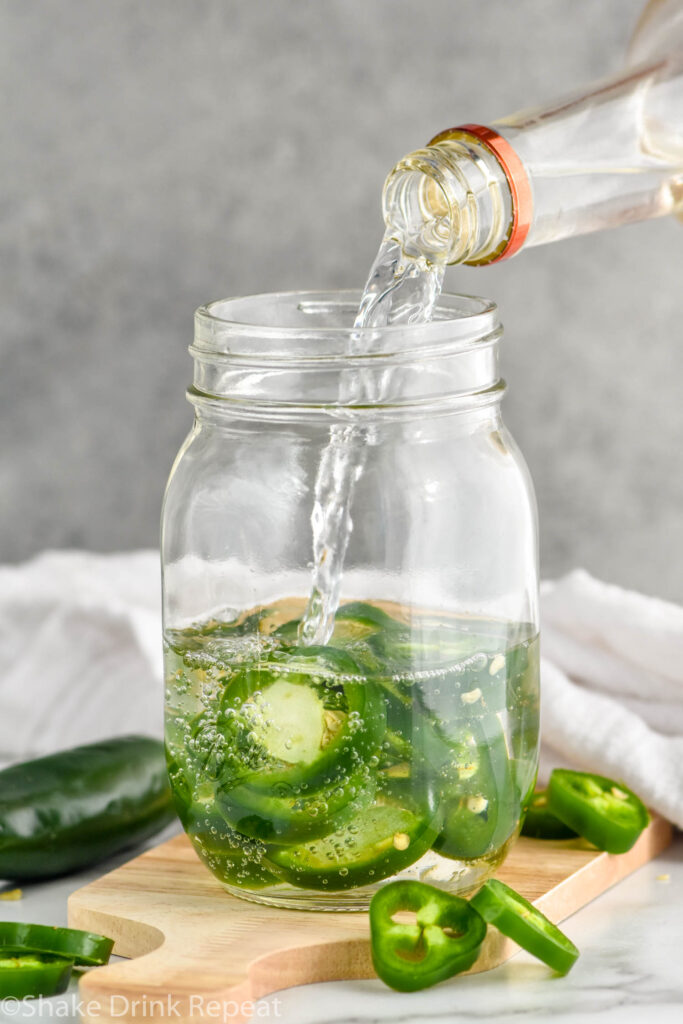 Photo of bottle of vodka being poured into jar with slices of jalapeno pepper for Jalapeno Infused Vodka recipe. Extra slices of jalapenos on counter beside jar.