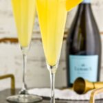 Photo of Mimosas garnished with orange slices. Bottle of Champagne in the background.