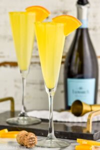 Photo of Mimosas garnished with orange slices. Bottle of Champagne in the background.