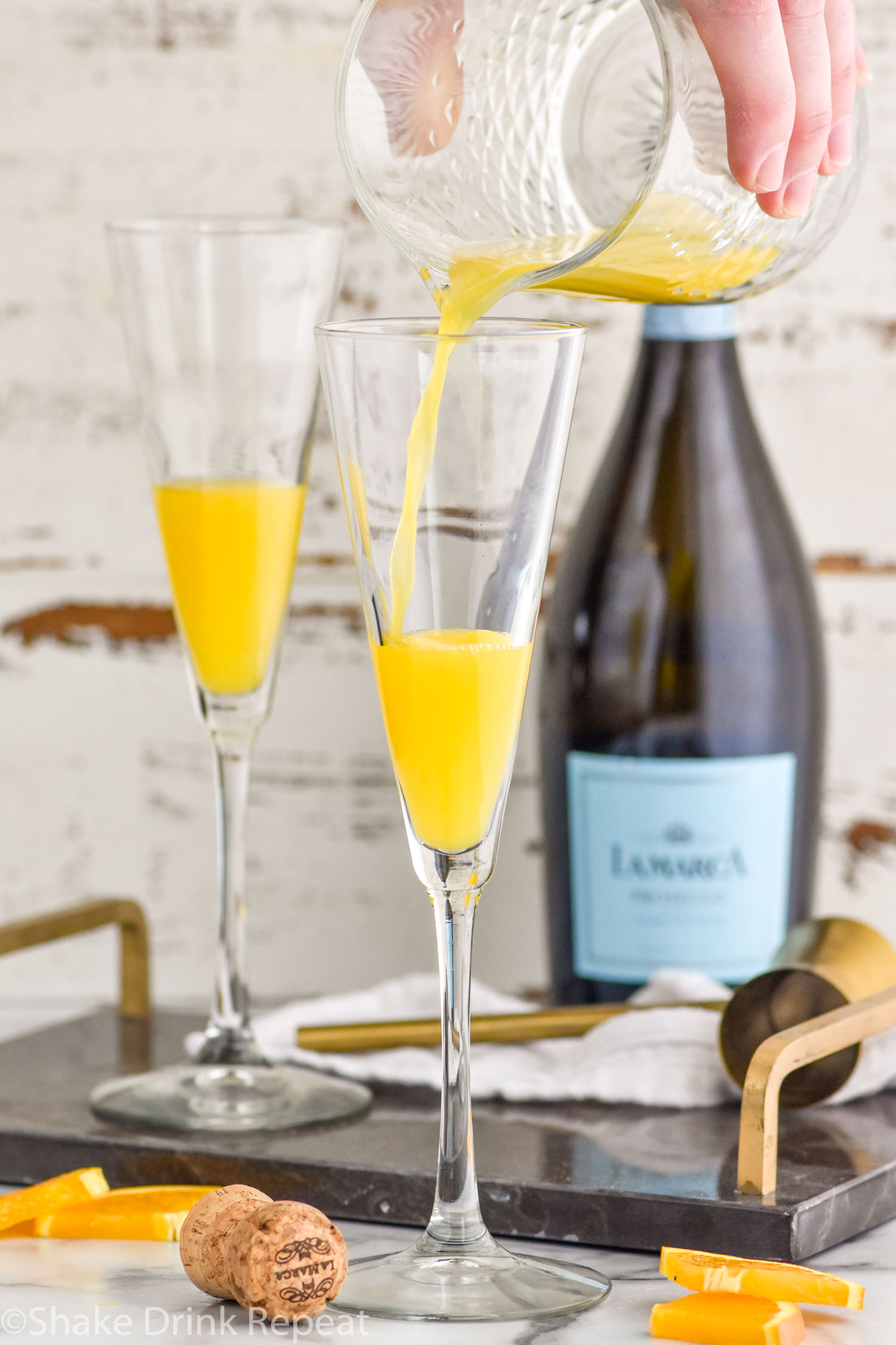 Photo of person's hand pouring orange juice into flute glasses for Mimosa recipe. Bottle of Champagne in the background for Mimosa recipe.
