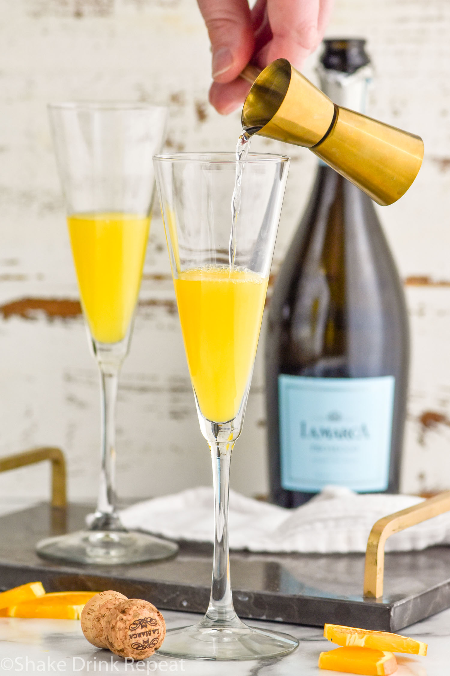 Photo of person's hand pouring Cointreau into flute glasses of orange juice for Mimosa recipe. Bottle of Champagne in background for Mimosa recipe.