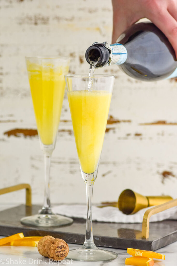 Photo of person's hand pouring Champagne into flute glasses of ingredients for Mimosa recipe.
