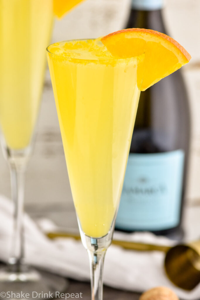 Close up photo of Mimosa garnished with orange slice. Bottle of Champagne in background.