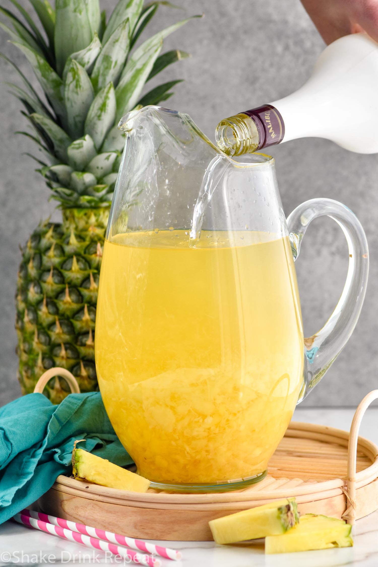 Photo of person's hand pouring Malibu rum into a pitcher of ingredients for Pina Colada Sangria recipe. A pineapple sits in the background.