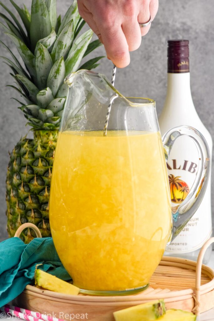 Photo of person's hand stirring pitcher of ingredients for Pina Colada Sangria. Pineapple and bottle of Malibu rum in background.