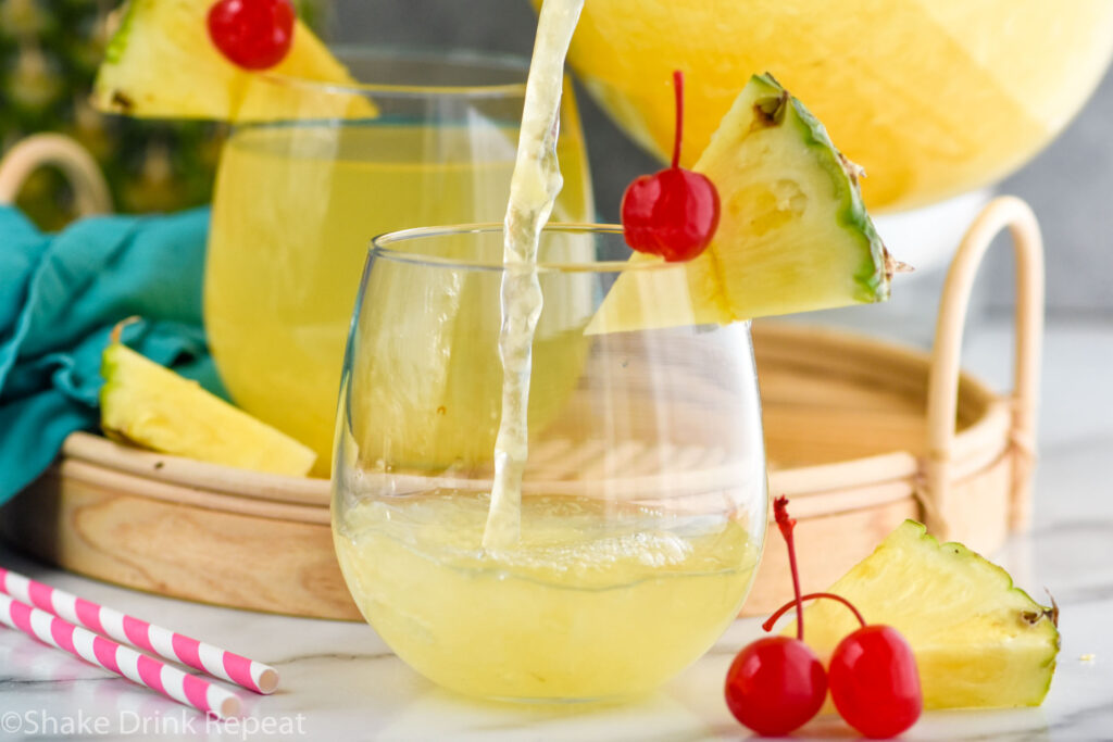 Photo of a pitcher of Pina Colada Sangria recipe being poured into a glass garnished with a pineapple wedge and a cherry.