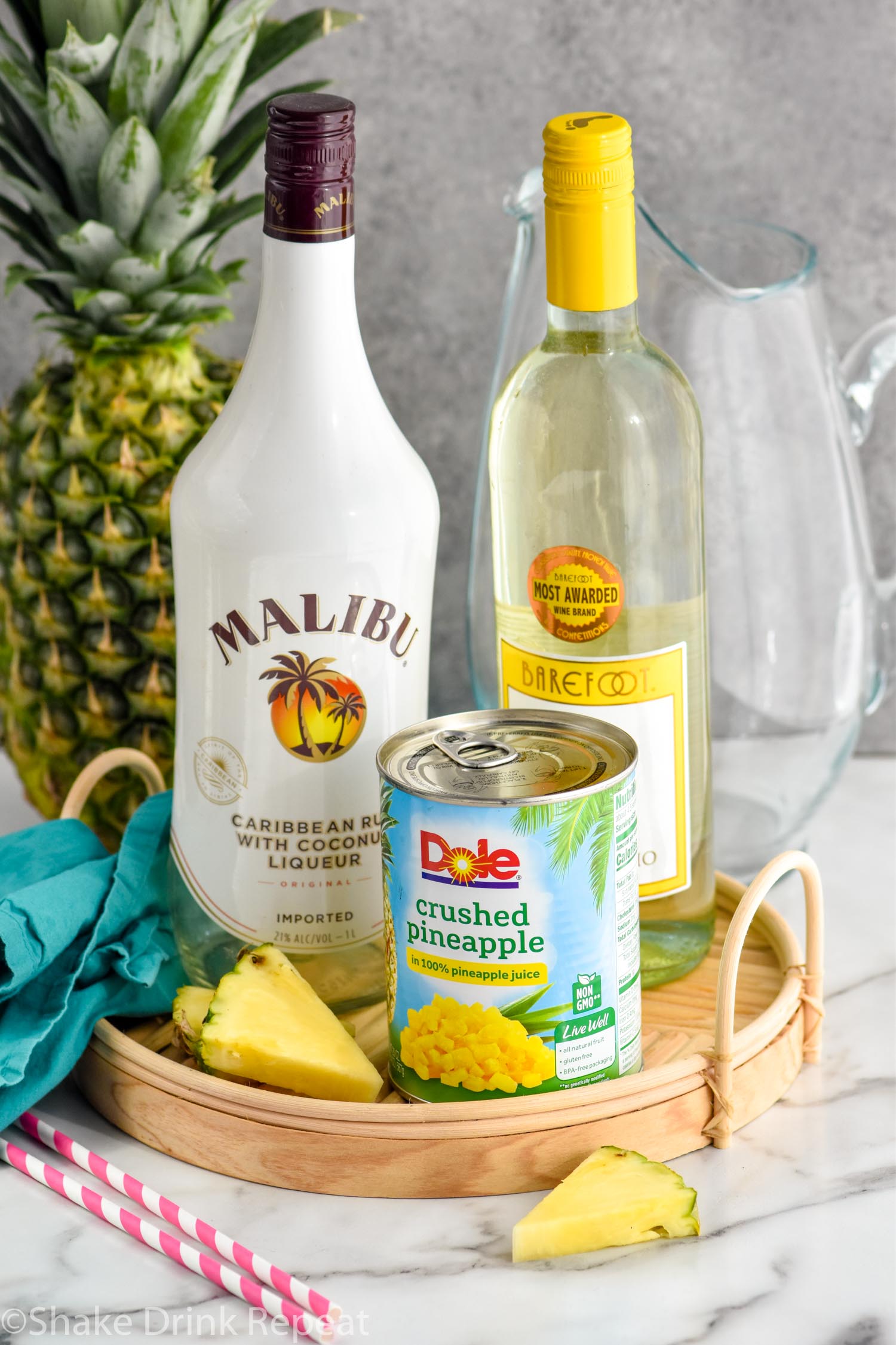 Bottle of Malibu rum, Moscato wine, and a can of crushed pineapple on platter for Pina Colada Sangria recipe. Glass pitcher and a pineapple in the background.