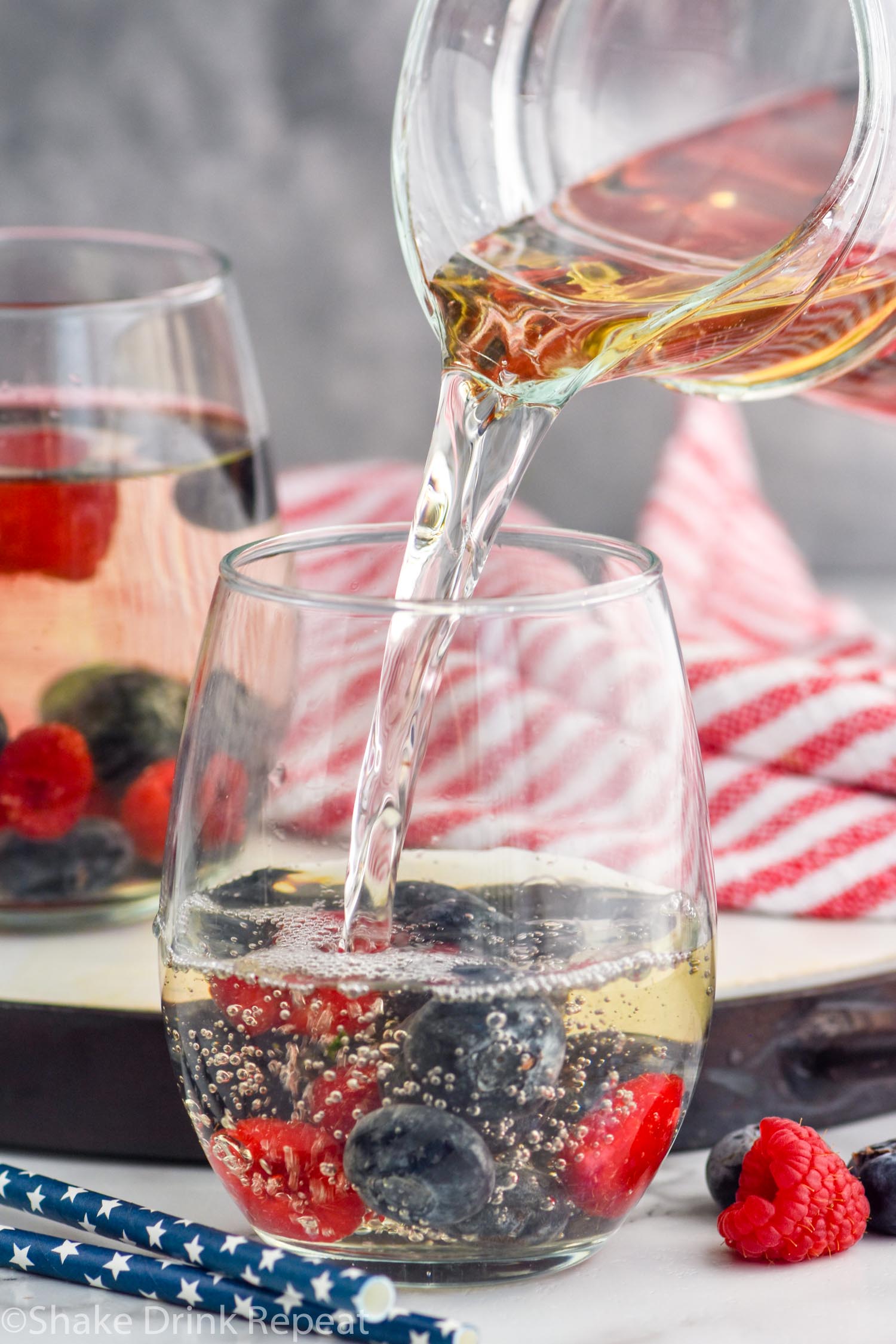 Side view of pitcher of Fourth of July Sangria being poured into a glass with blueberries and raspberries. Another full glass in the background, extra fruit beside glasses.