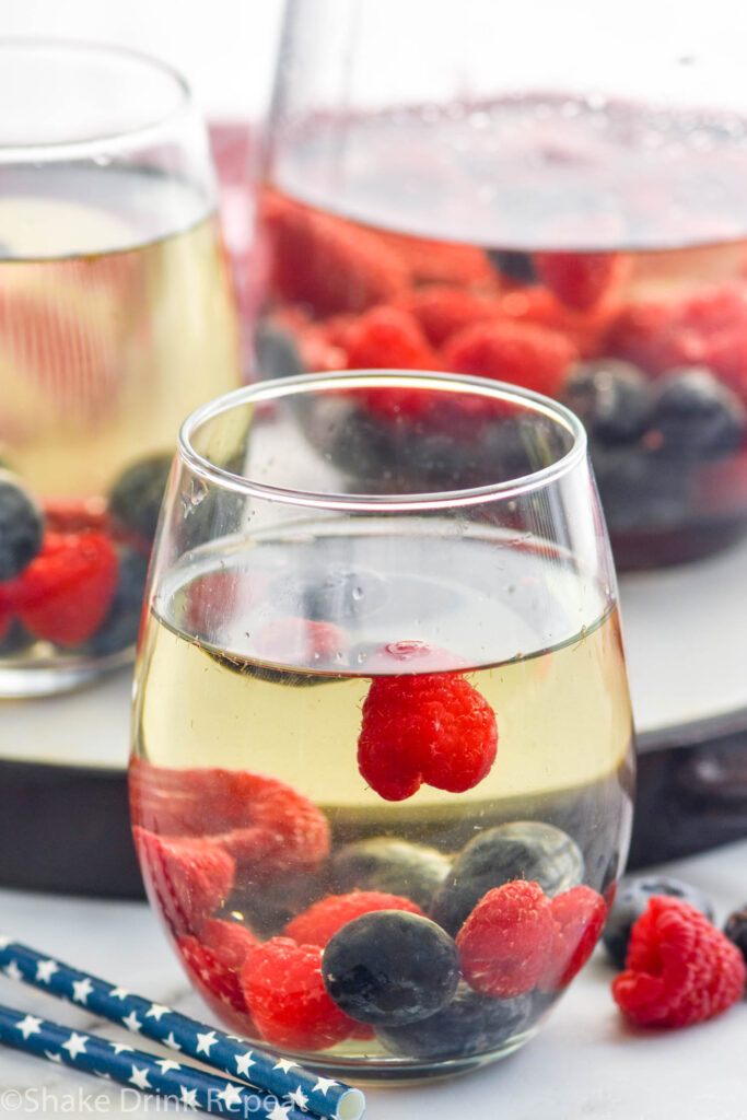 Side view of a glass of Fourth of July Sangria garnished with raspberries and blueberries. More glasses in the background, extra fruit beside glass.