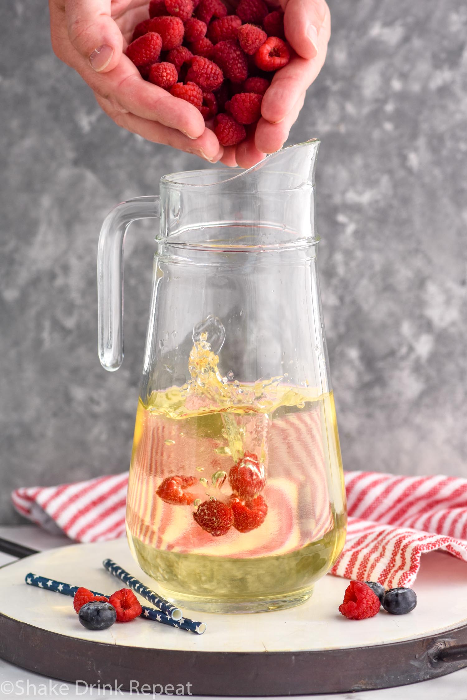 Side view of person's hand dropping raspberries into pitcher of ingredients for Fourth of July Sangria recipe. Extra blueberries and raspberries on counter beside pitcher.