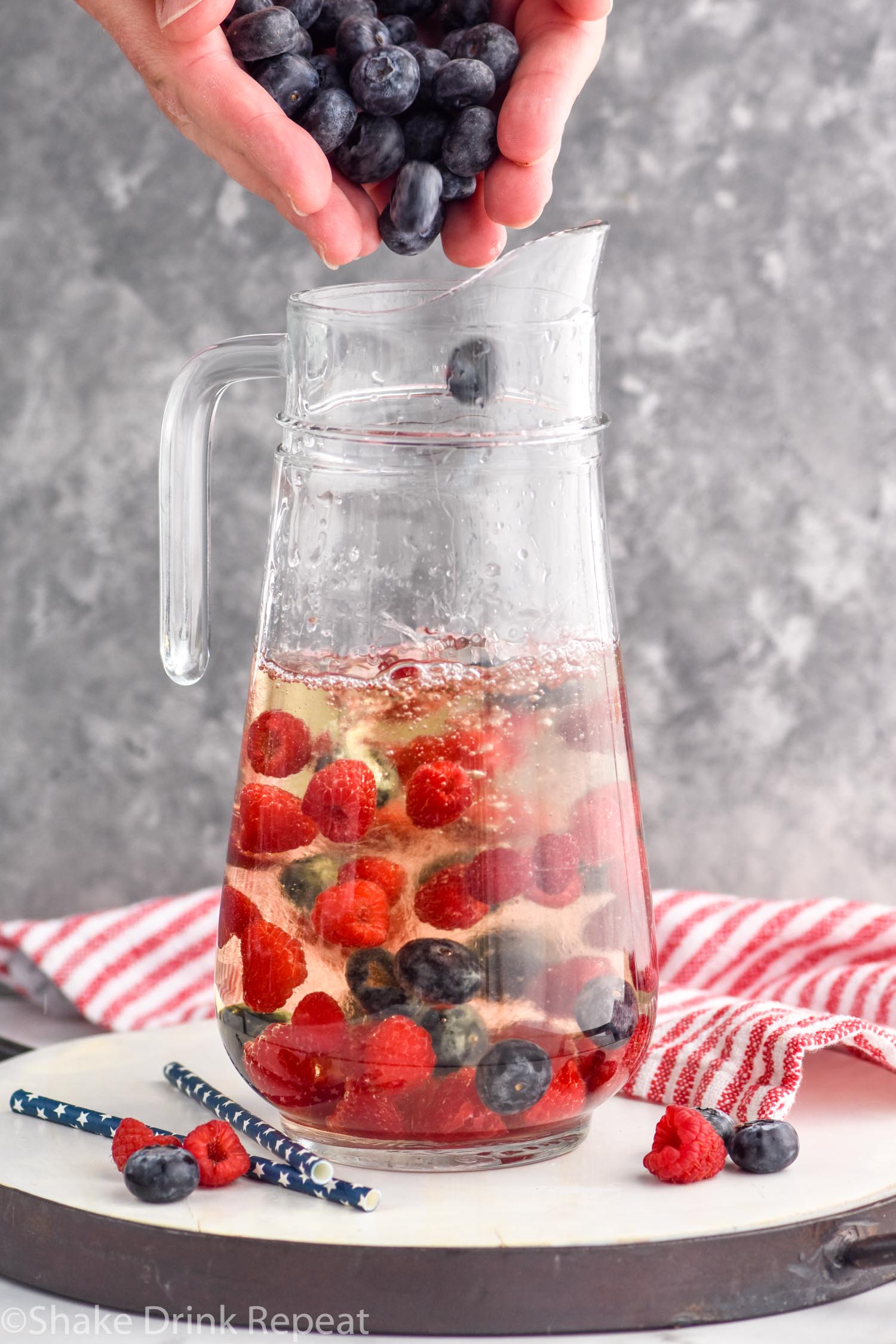 Side view of person's hand dropping blueberries into pitcher of Fourth of July Sangria recipe. Extra raspberries and blueberries beside pitcher.