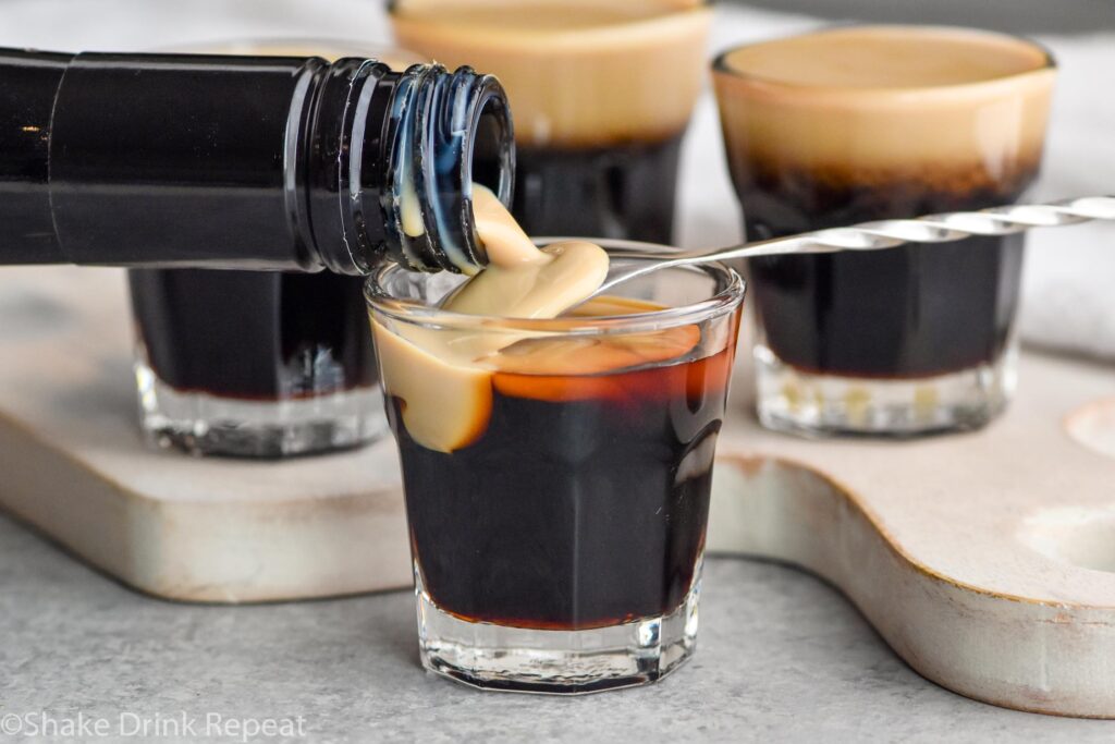 Side view of Bailey's being poured over the back of a spoon into a shot glass of Kahlua for Baby Guinness Shot recipe.