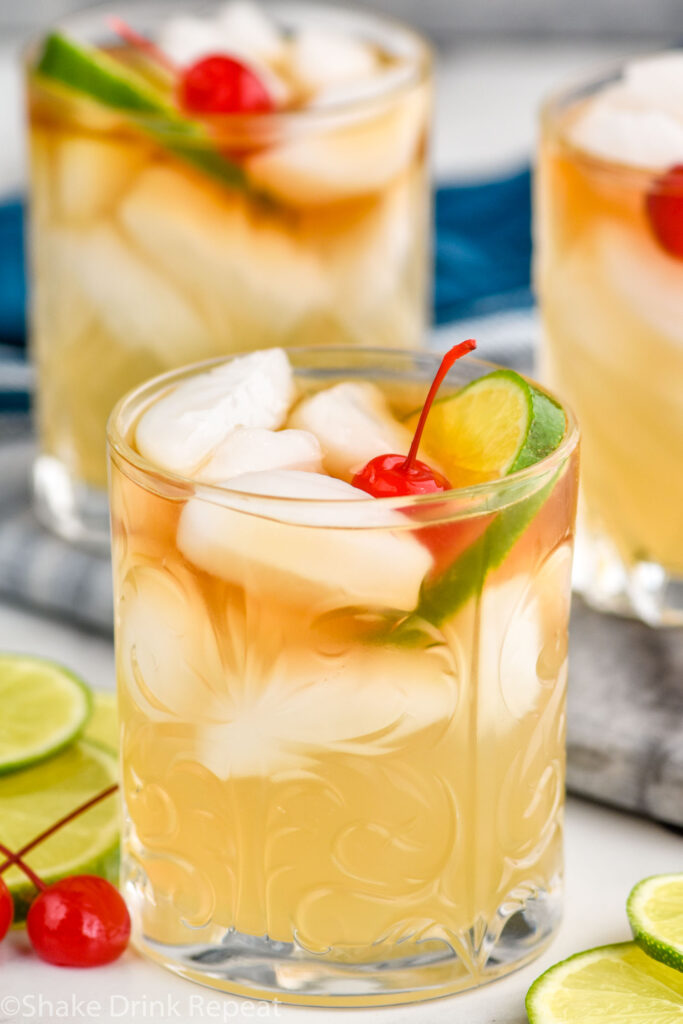 Side view of Mai Tai cocktail garnished with lime wedge and cherry. More glasses of Mai Tai in the background. Lime wedges and cherries beside the glasses for garnish.