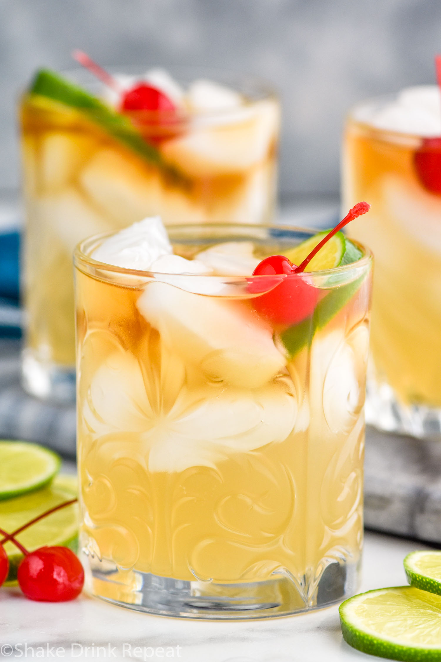 Side view of Mai Tai cocktail garnished with lime wedge and cherry. More glasses of Mai Tai in the background. Lime wedges and cherries beside the glasses for garnish.