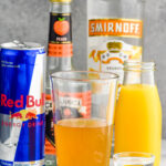 Side view of a pint glass and a shot glass of ingredients for cactus cooler shot recipe. Bottle of orange juice, peach schnapps, mandarin vodka, and a can of red bull.