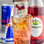 Side view of a cherry bomb shot with the shot glass splashing into the pint glass. Can of red bull, bottle of cherry vodka, and bottle of grenadine behind glass.