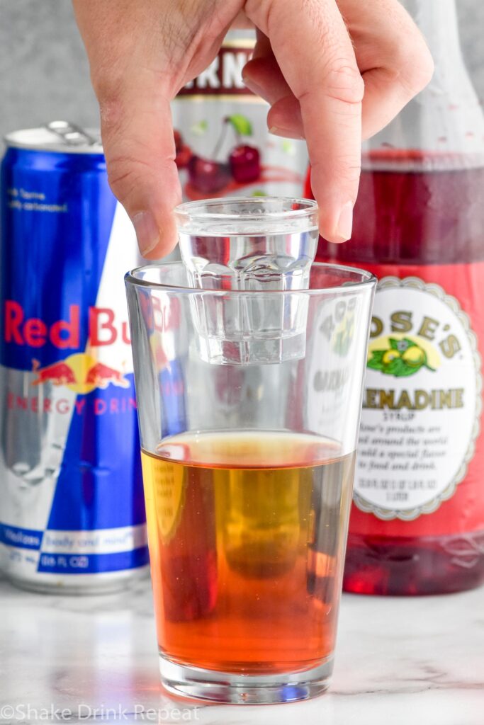 Side view of person's hand holding a shot glass of cherry vodka into a pint glass of red bull and grenadine for Cherry Bomb Shot recipe. Can of red bull, bottle of grenadine, and bottle of cherry vodka on the counter behind glass.