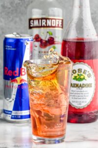 Side view of a cherry bomb shot with the shot glass splashing into the pint glass. Can of red bull, bottle of cherry vodka, and bottle of grenadine behind glass.