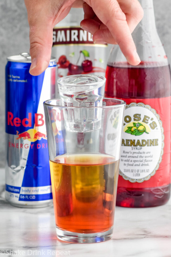 Side view of person's hand dropping a shot glass of cherry vodka into a pint glass of red bull and grenadine for Cherry Bomb Shot recipe. Can of red bull, bottle of grenadine, and bottle of cherry vodka on the counter behind glass.