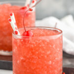 Side view of glasses of Dirty Shirley Slush garnished with cherries and straws. Extra cherries beside glasses.