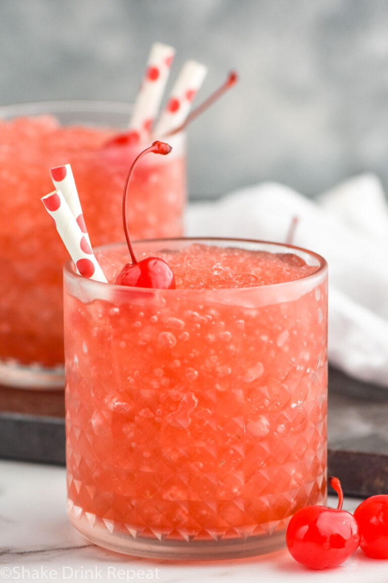 Side view of glasses of Dirty Shirley Slush garnished with cherries and straws. Extra cherries beside glasses.