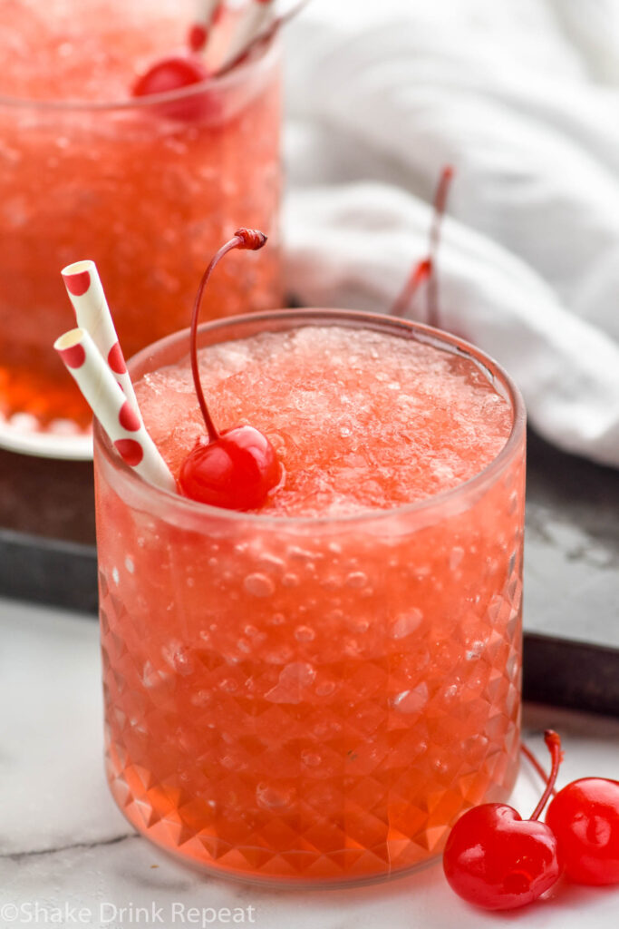 Side view of a glass of Dirty Shirley Slush garnished with a cherry and straws. Another glass in the background. Cherries beside glass.