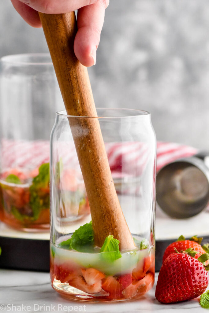 Side view of person's hand muddling ingredients for Strawberry Mojito recipe. Another glass of ingredients in the background. Strawberries on the counter beside glasses for garnish.