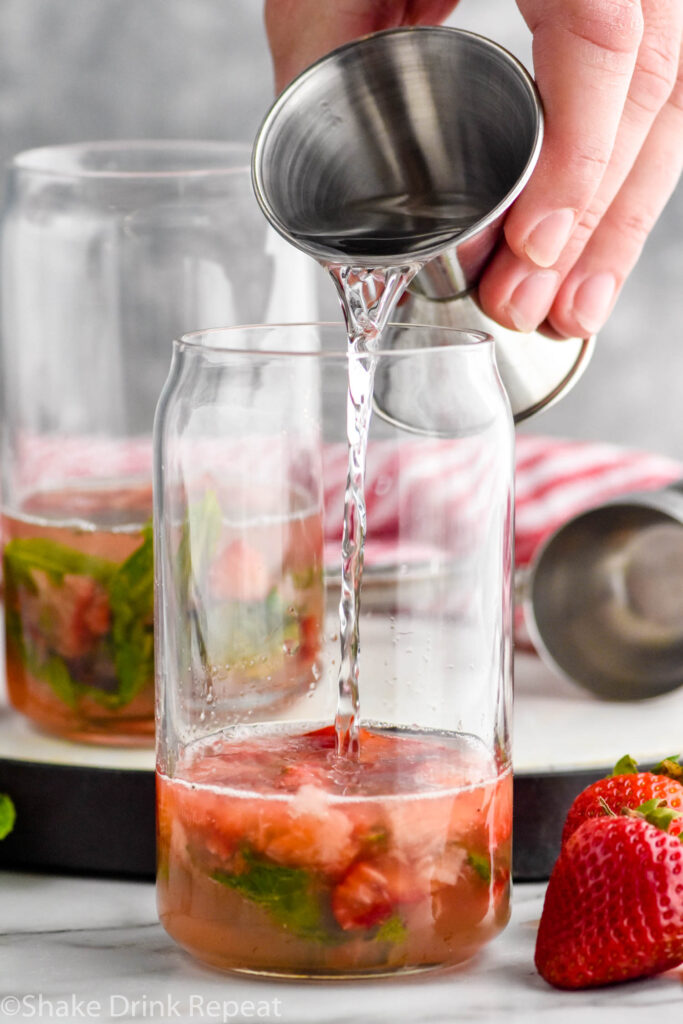 Side view of person's hand pouring rum into a glass of ingredients for Strawberry Mojito recipe. Another glass of ingredients behind. Strawberries for garnish on counter beside glass.