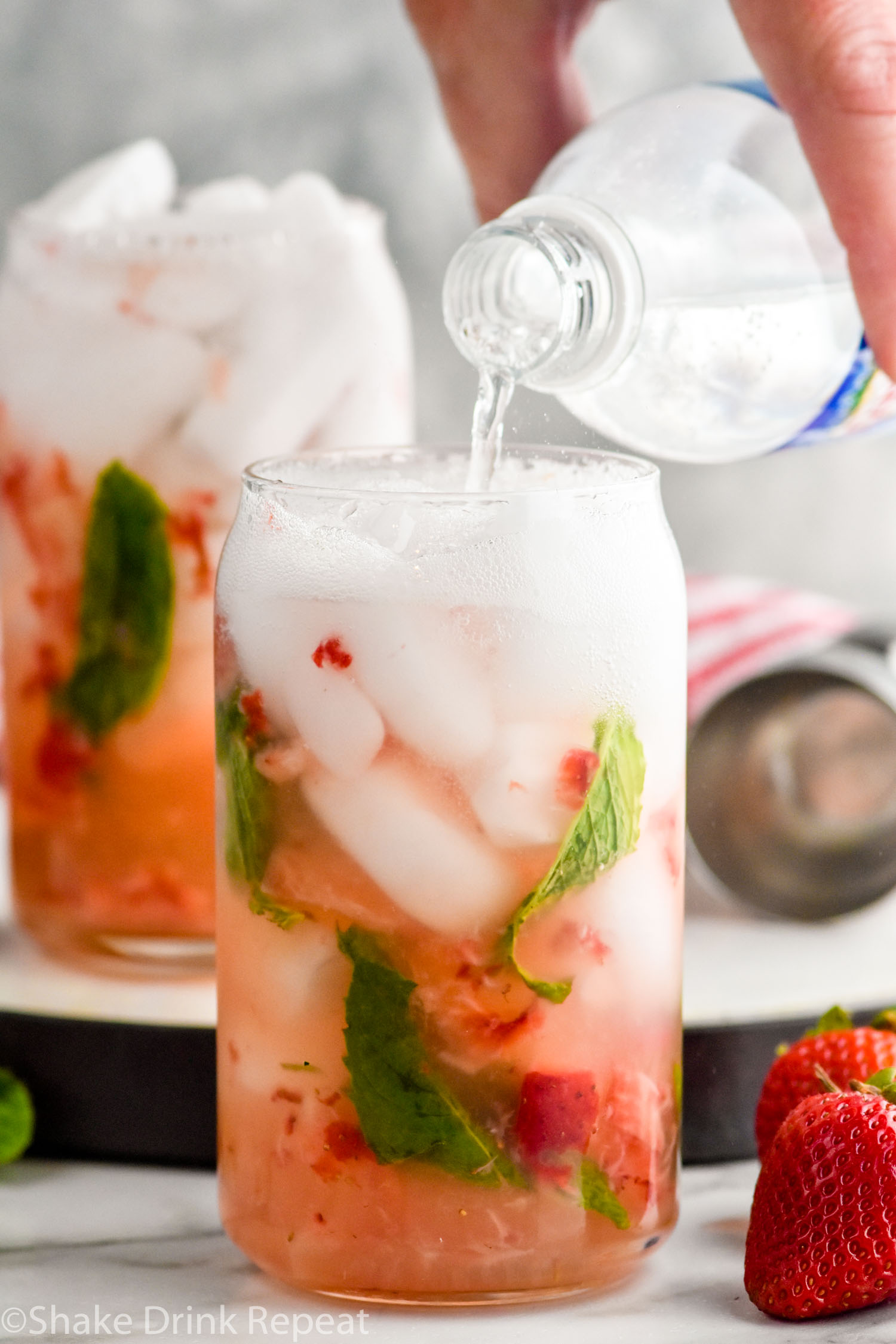 Side view of person's hand pouring club soda into glass of ingredients for Strawberry Mojito recipe. Another glass of Strawberry Mojito in the background. Strawberries beside glasses.