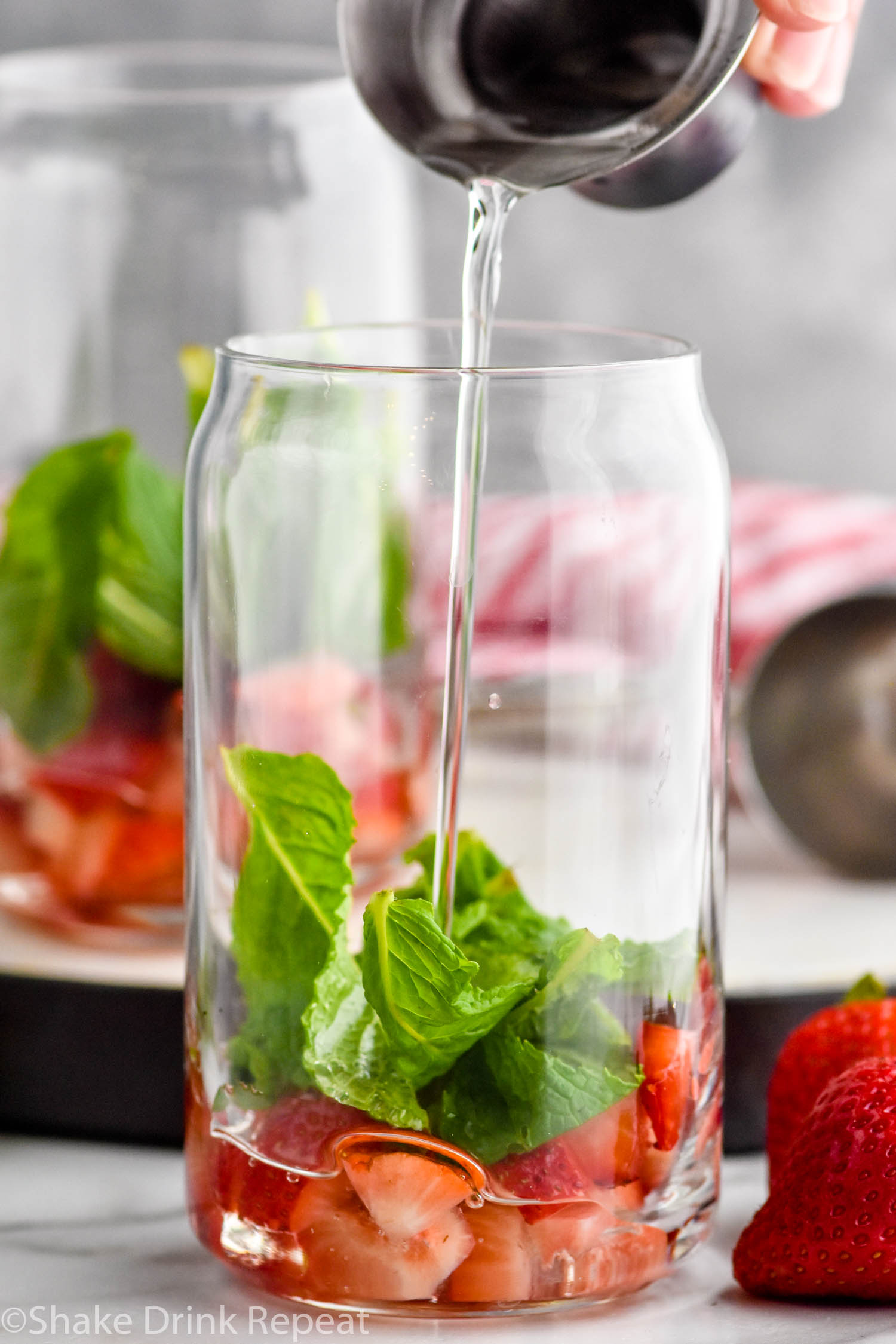 Side view of person's hand pouring simple syrup into glass of ingredients for Strawberry Mojito recipe. Another glass of ingredients in the background. Strawberries beside glasses.
