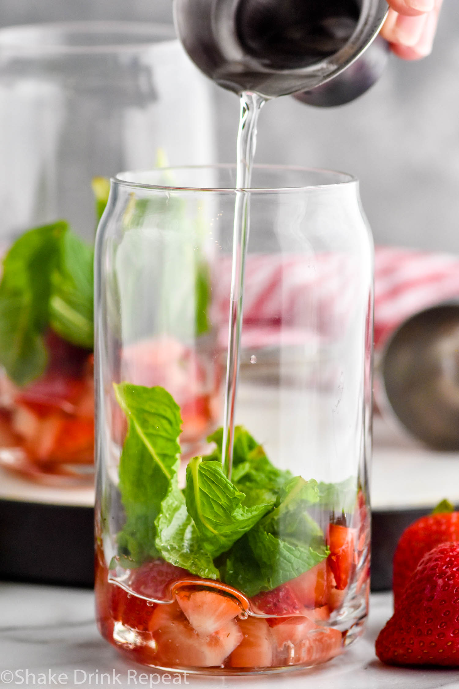 Side view of person's hand pouring simple syrup into glass of ingredients for Strawberry Mojito recipe. Another glass of ingredients in the background. Strawberries beside glasses.
