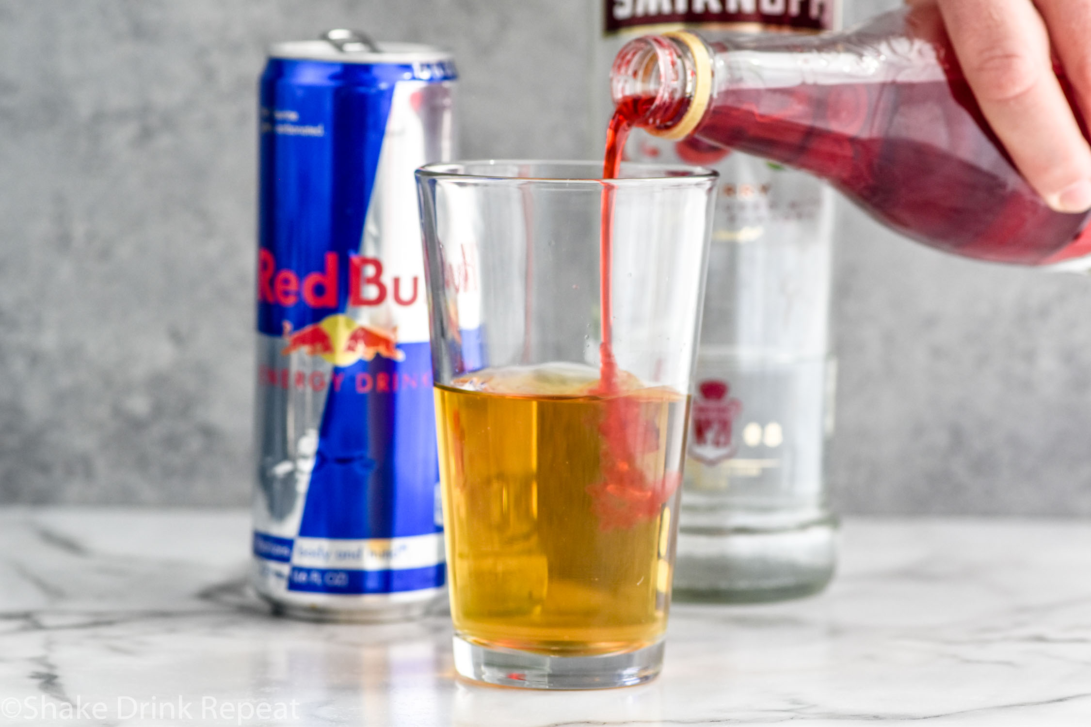 Side view of person's hand pouring grenadine into a pint glass with Red Bull for Cherry Bomb Shot recipe. Can of Red Bull and bottle of cherry vodka in the background.