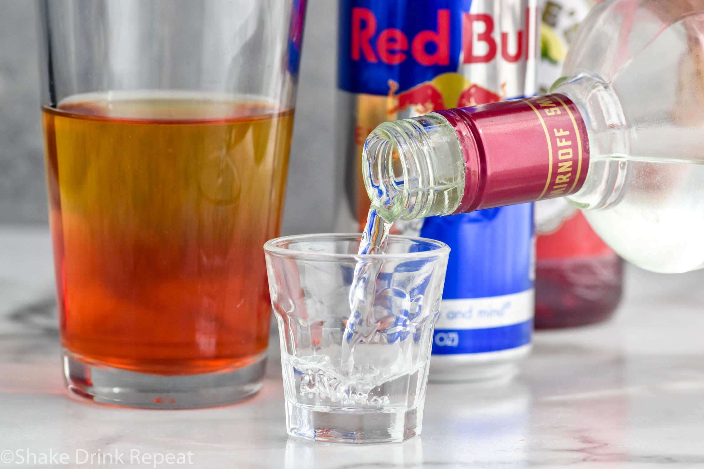 Side view of bottle of cherry vodka being poured into a shot glass for Cherry Bomb Shot recipe. Pint glass of ingredients for Cherry Bomb Shot and can of Red Bull on counter.