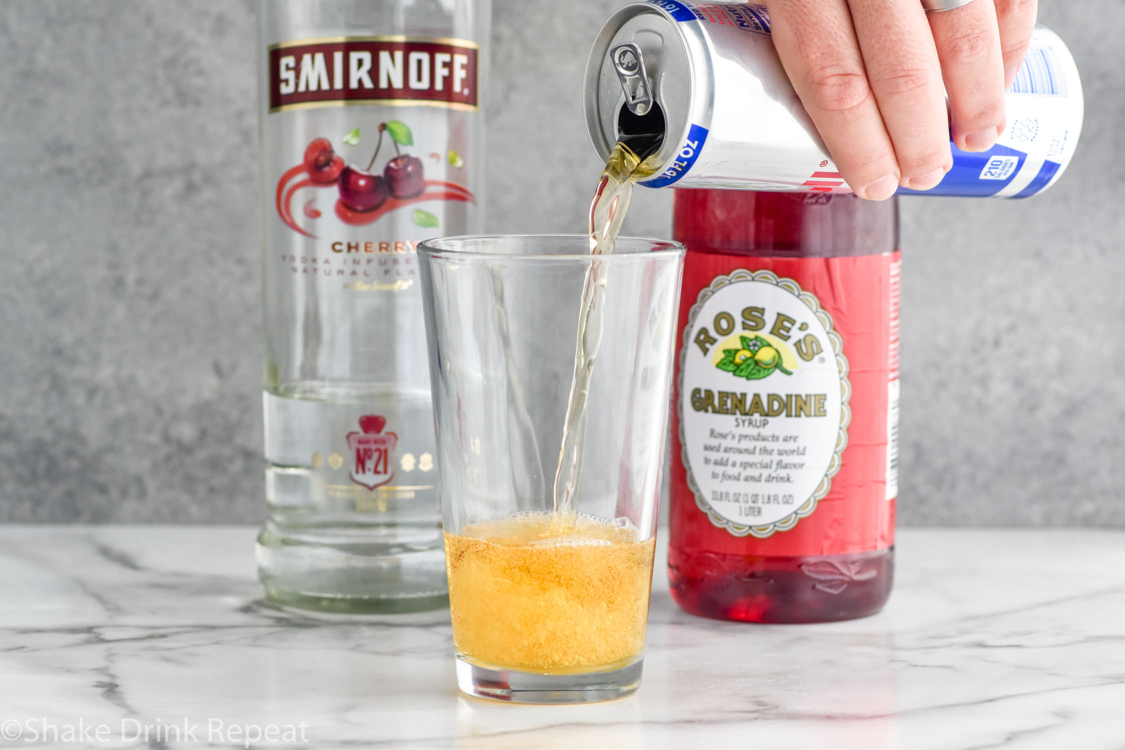 Side view of person's hand pouring a can of Red Bull into a pint glass for Cherry Bomb Shot recipe. Bottles of cherry vodka and grenadine behind glass for recipe.