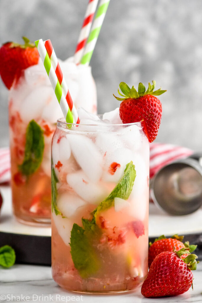 Side view of glass of strawberry mojito garnished with a strawberry on the rim and straws. Another strawberry mojito in the background. Strawberries on counter beside glass.