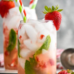 Side view of glass of strawberry mojito garnished with a strawberry on the rim and straws. Another strawberry mojito in the background. Strawberries on counter beside glass.