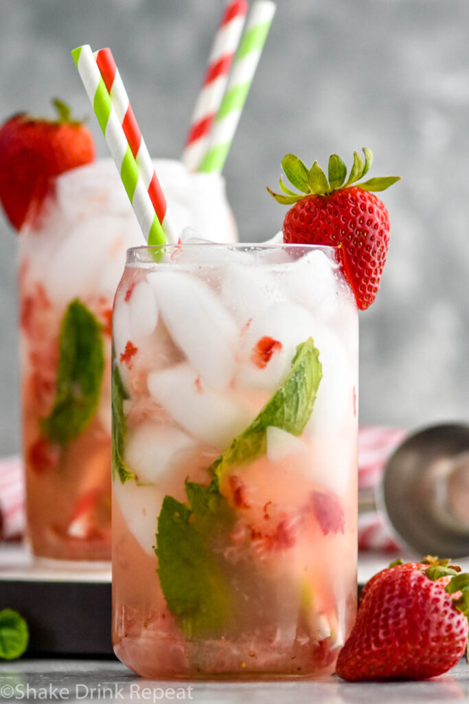 Side view of glass of strawberry mojito garnished with a strawberry on the rim and straws. Another strawberry mojito in the background. Strawberries on counter beside glass.