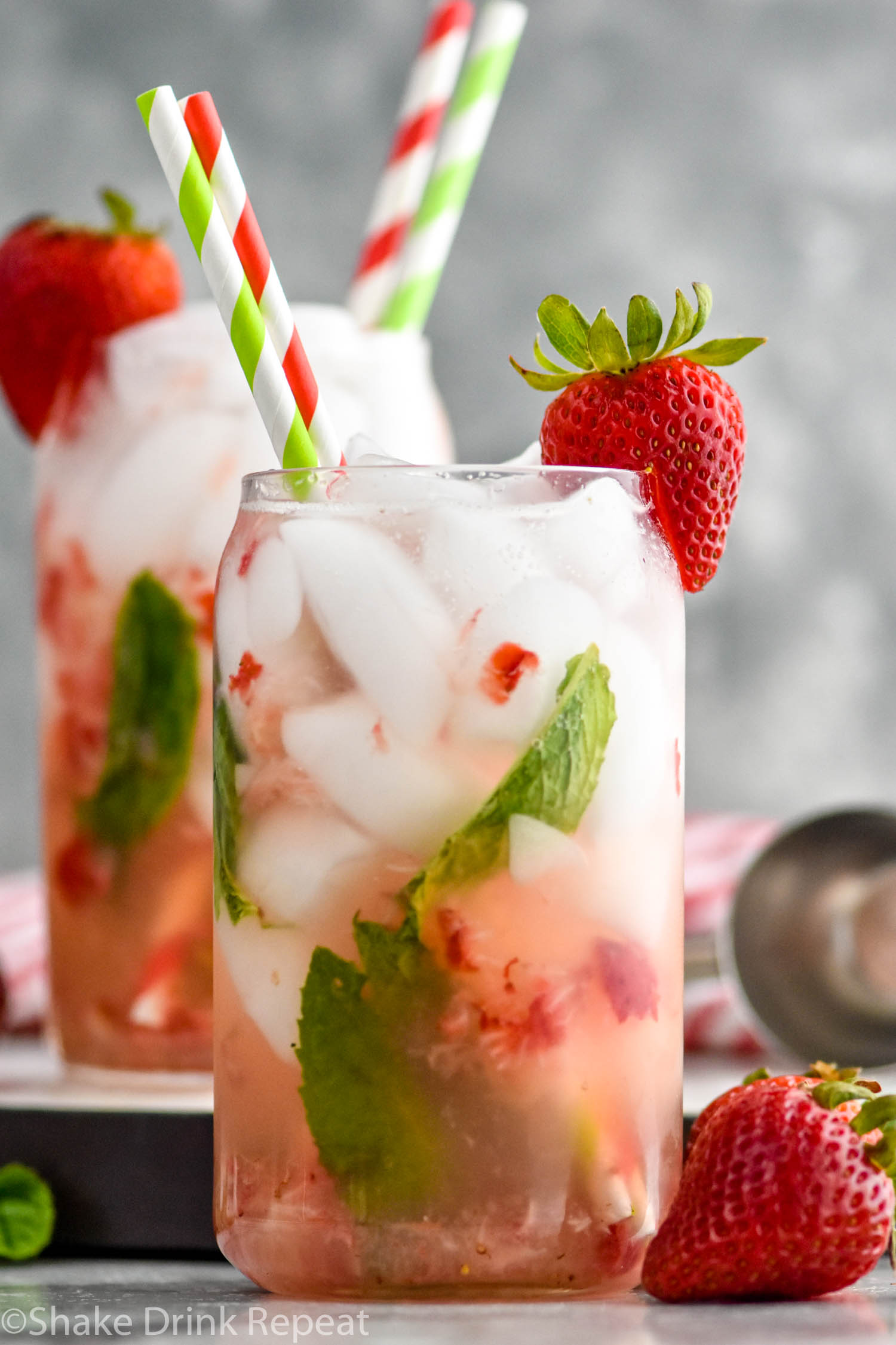 Side view of glass of strawberry mojito garnished with a strawberry on the rim and straws. Another strawberry mojito in the background. Strawberries on counter beside glass.