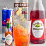 Photo of shot glass splashing into pint glass of energy drink for Chuck Norris Shot recipe. hot sauce, red bull, cherry vodka, and grenadine in the background.