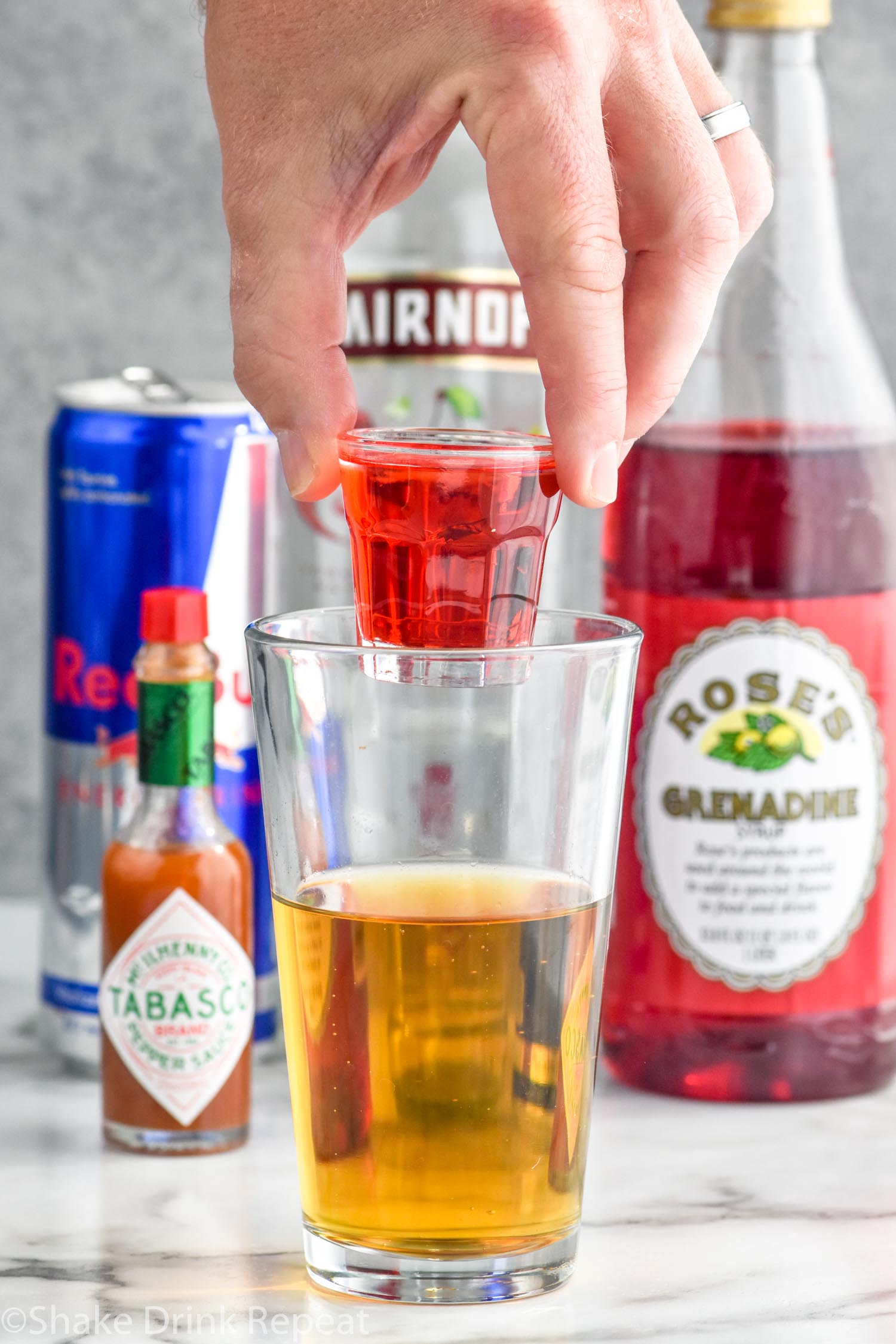 Photo of person's hand holding a shot glass of ingredients over a pint glass of energy drink for Chuck Norris Shot recipe. Hot sauce, red bull, cherry vodka, and grenadine in the background.