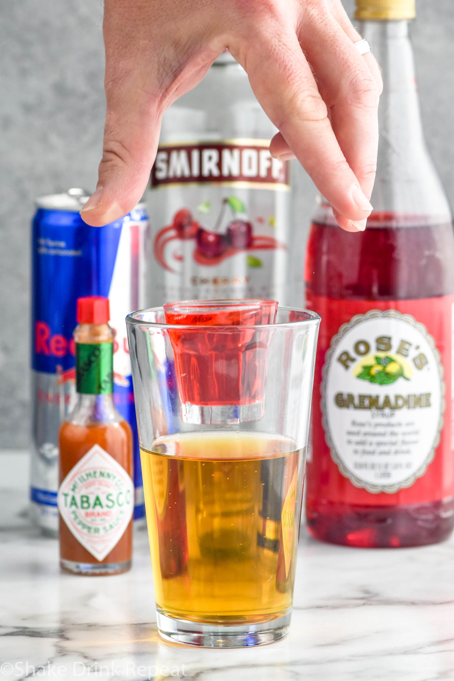 Photo of person's hand dropping a shot glass of ingredients into a pint glass of energy drink for Chuck Norris Shot recipe. Hot sauce, red bull, cherry vodka, and grenadine behind Chuck Norris Shot.