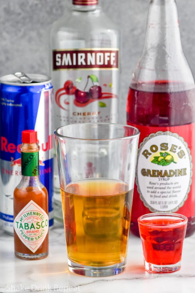 Side view of ingredients for Chuck Norris Shot, including hot sauce, a glass of Red Bull, a shot glass of vodka and grenadine, a can of red bull, a bottle of cherry vodka, and a bottle of grenadine.