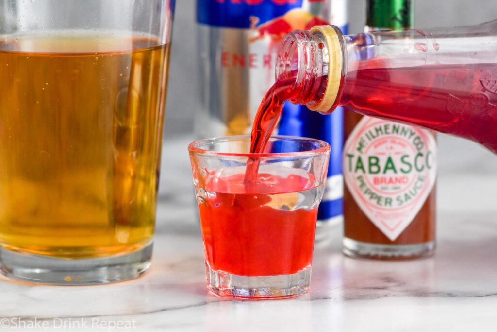 Side view of grenadine being poured into shot glass for Chuck Norris Shot recipe. Glass of red bull, red bull can, and hot sauce behind shot glass.