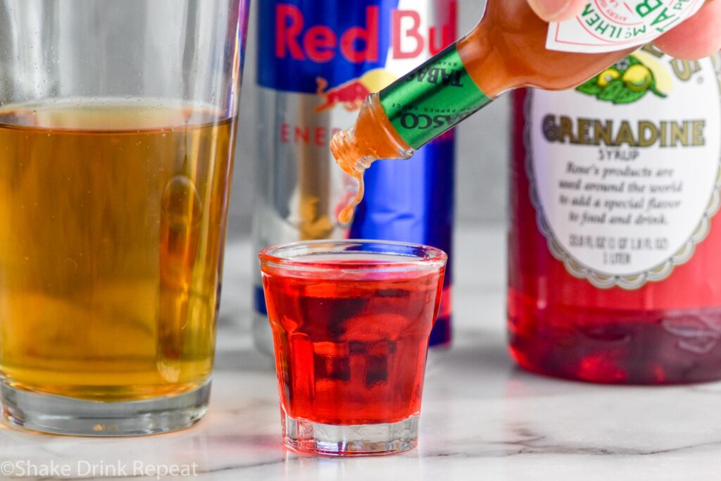 Side view of person's hand dashing hot sauce into shot glass of ingredients for Chuck Norris Shot recipe. Glass of red bull, red bull can, and bottle of grenadine behind shot glass.