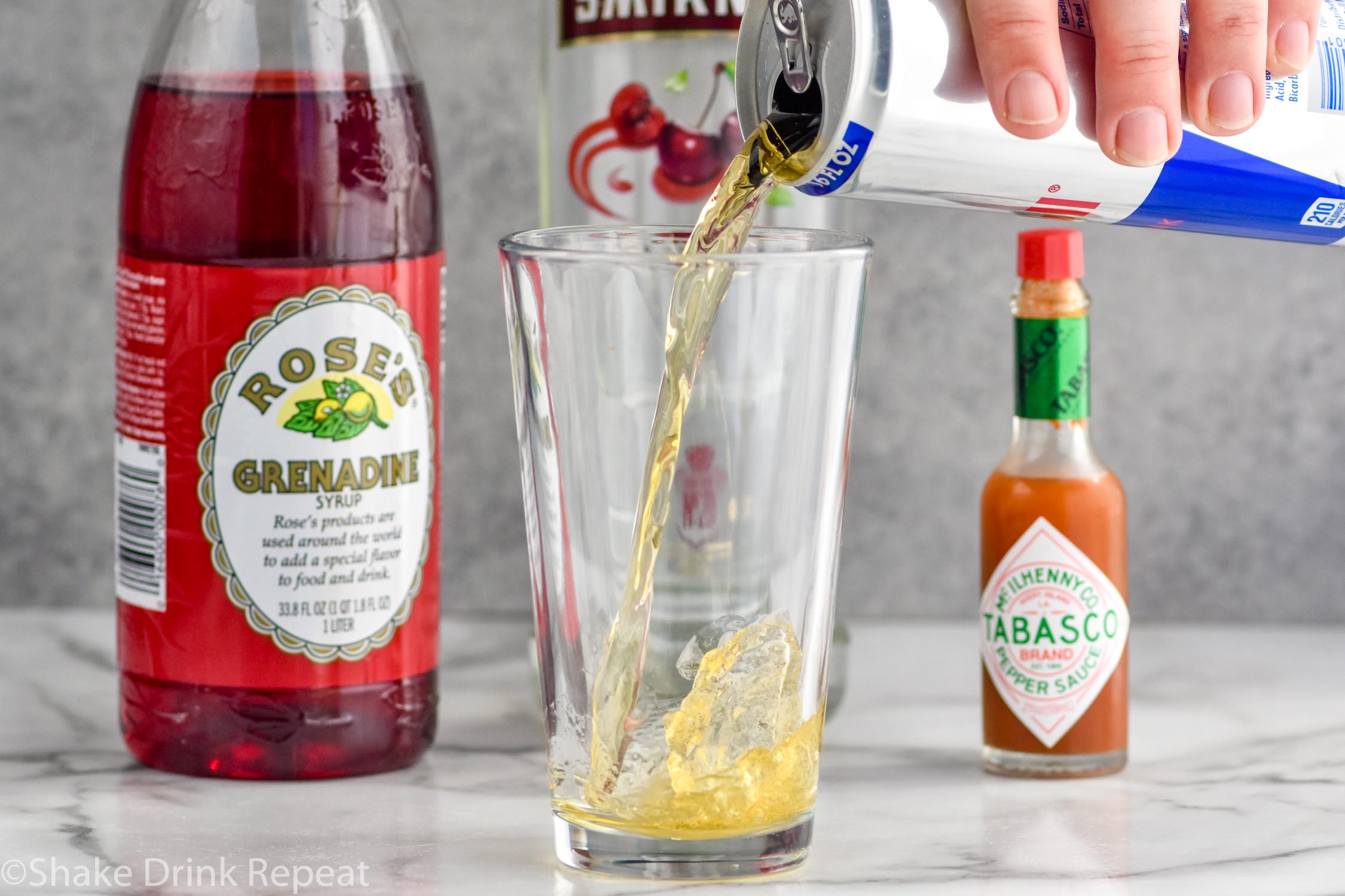 Side view of person's hand pouring red bull into a shot glass for Chuck Norris Shot recipe. Grenadine, cherry vodka, and hot sauce behind glass also for recipe.