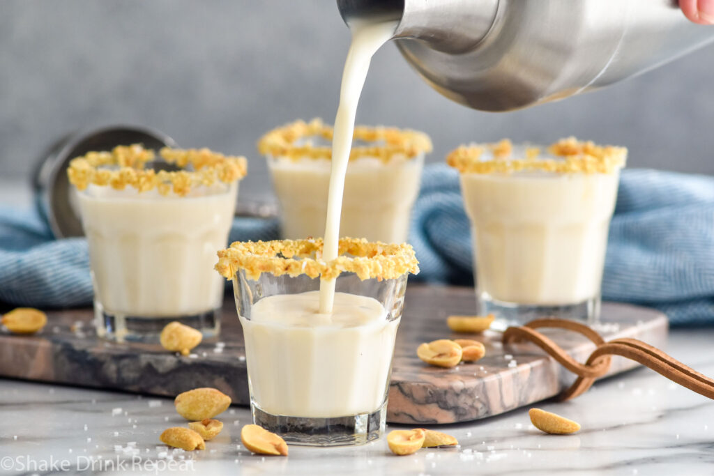 Side view of cocktail shaker of Salted Nut Roll shot recipe being poured into prepared shot glasses with crushed peanuts. Three more Salted Nut Roll shots in the background, extra peanuts on salt on the counter.