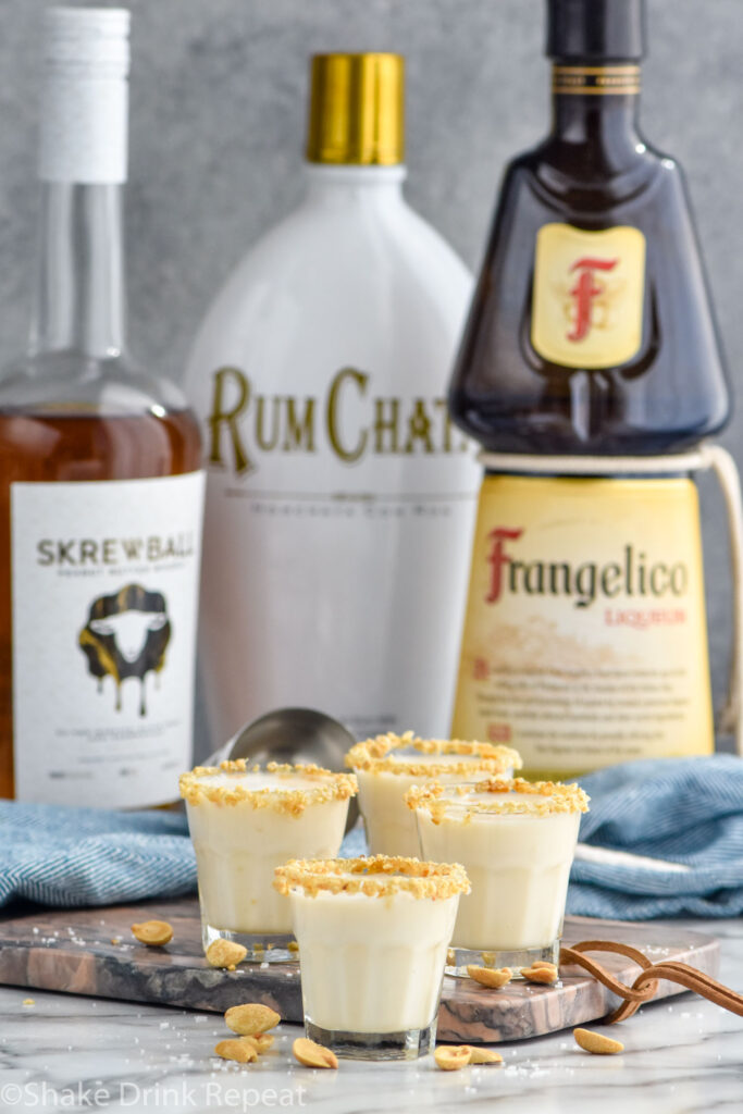 Side view of Salted Nut Roll shots garnished with crushed peanuts on the rim. Bottles of Skrewball peanut butter whiskey, RumChata, and Frangelico behind shots.